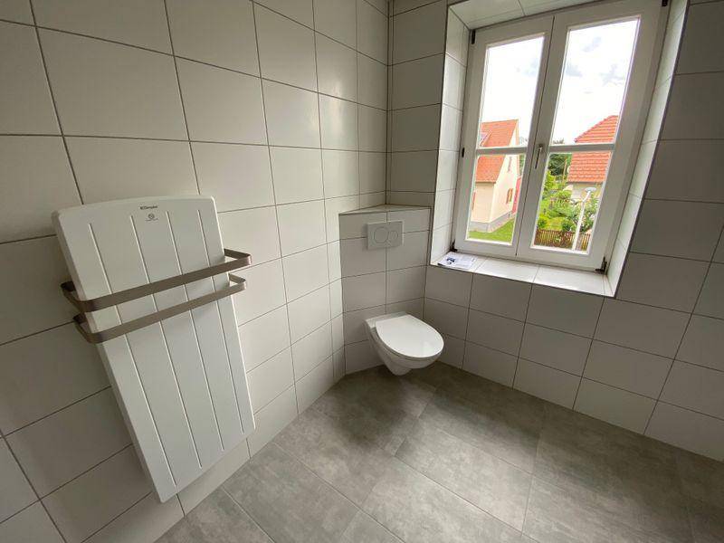 Helles Badezimmer mit Wand-WC, Heizkörper und Fenster, komplett gefliest in Weiß und Grau.