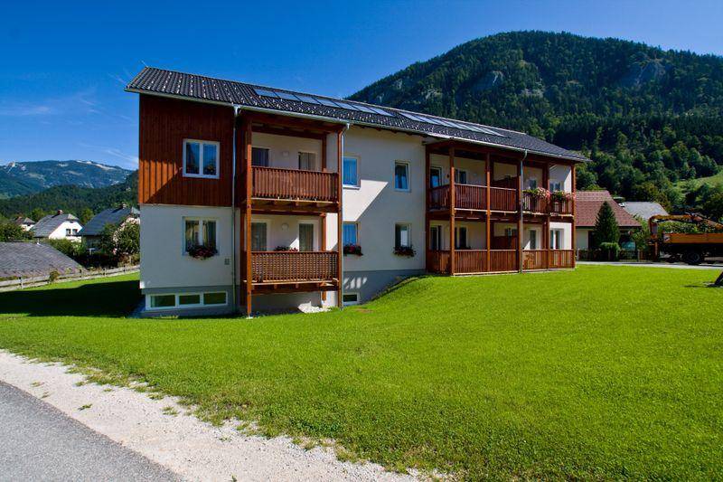 Ansicht des Gebäudes mit Holzfassade, Balkonen und Solarpaneelen auf dem Dach, umgeben von grüner Natur.