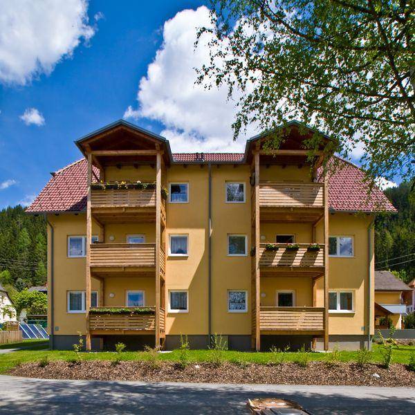 Vorderansicht eines modernen Gebäudes mit gelber Fassade, mehreren Holzbalkonen und rotem Ziegeldach unter blauem Himmel.