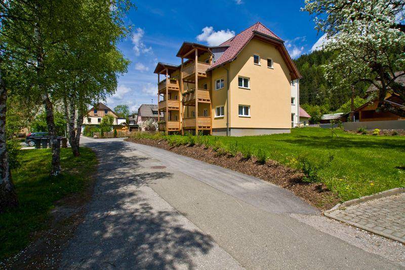 Seitenansicht des Gebäudes mit gelber Fassade, Holzbalkonen und Blick auf die umliegende grüne Landschaft und Berge.