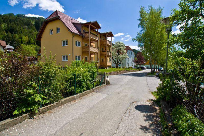 Gebäudeansicht von der Straße aus, umgeben von Bäumen und grüner Natur, mit Blick auf die Berge im Hintergrund.