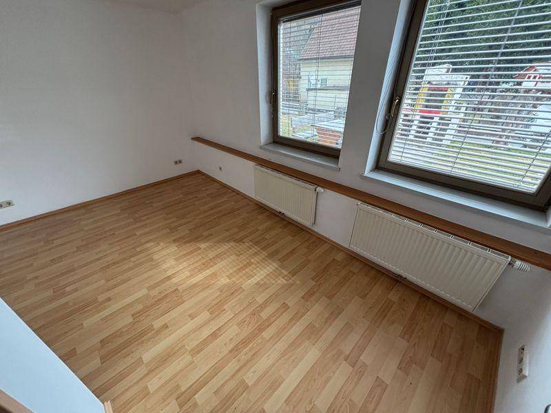 Helles Zimmer mit Laminatboden und zwei Fenstern, ausgestattet mit Jalousien.