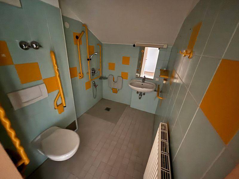 Barrierefreies Badezimmer mit gelben Haltegriffen, Dusche, WC und Waschbecken in hellblauen Fliesen.