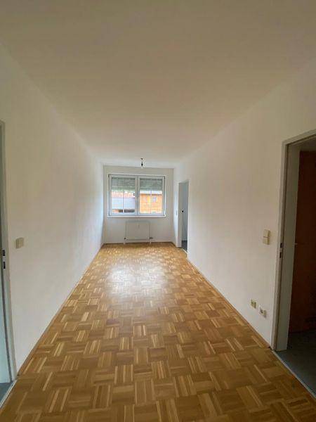 Heller, leerer Raum mit Fischgrätparkettboden und einem Fenster mit Rollladen.