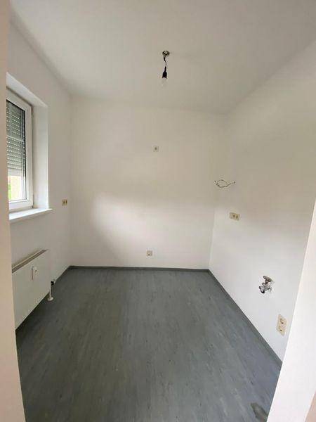 Leerer Raum mit grauem Vinylboden, Fenster und Anschlüssen für eine zukünftige Küche.
