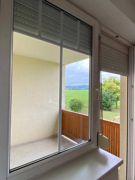 Blick aus einem Fenster auf einen Balkon und eine weite grüne Landschaft mit Hügeln.