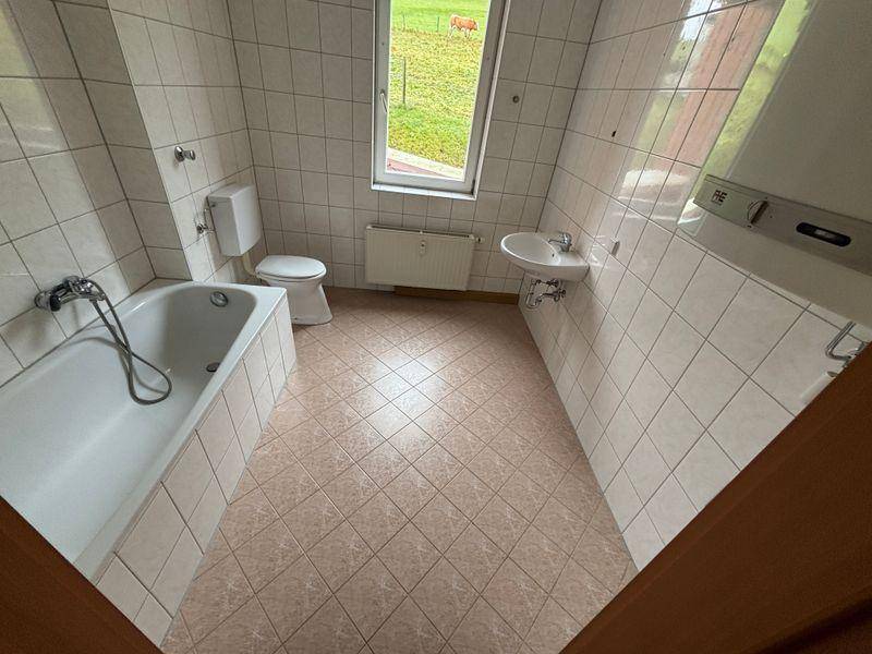 Funktionales Badezimmer mit Badewanne, WC, Waschbecken und Fenster mit Blick ins Grüne.