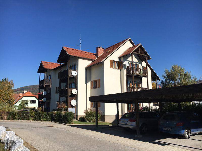 Mehrfamilienhaus mit mehreren Balkonen und Carport-Stellplätzen unter blauem Himmel.