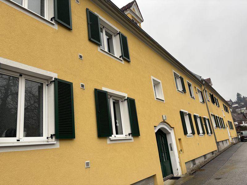 Nahaufnahme der gelben Gebäudefassade mit grünen Fensterläden und weiß gerahmten Fenstern.