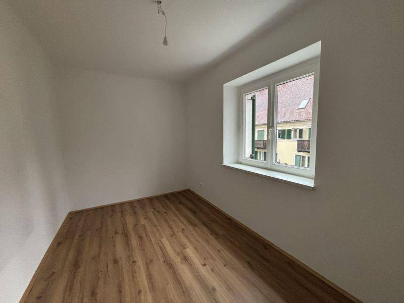 Helles Zimmer mit Laminatboden und Fenster, das auf andere Gebäude blickt.