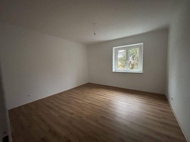 Großes, helles Zimmer mit Laminatboden und einem Fenster mit Blick ins Grüne.