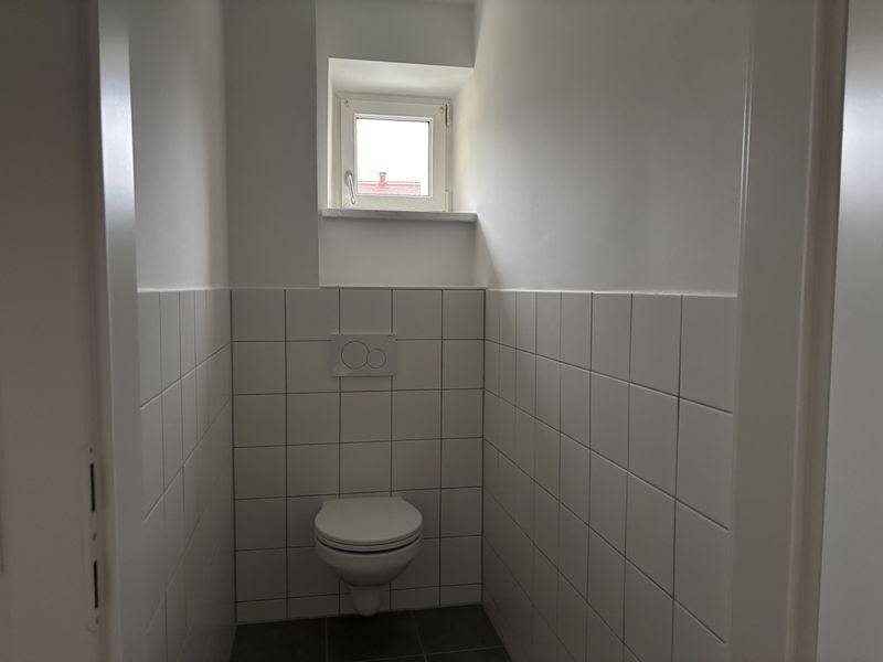 Separates WC mit wandhängender Toilette, weißen Fliesen und kleinem Fenster.
