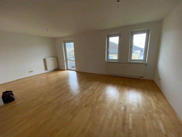 Geräumiges, leeres Wohnzimmer mit hellem Parkettboden und Zugang zu einem möglichen Balkon.