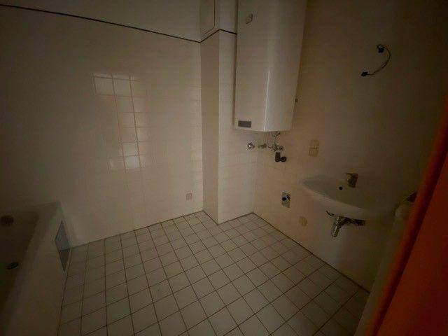 Badezimmer mit Fliesenboden, Waschbecken und einem Warmwasserboiler, Badewanne teilweise sichtbar.