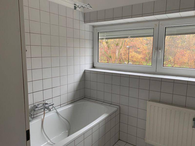 Helles Badezimmer mit weißen Fliesen, einer Badewanne und einem Fenster mit Blick ins Grüne.