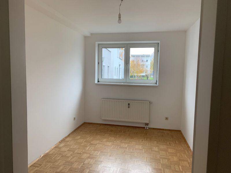 Leeres Zimmer mit Parkettboden, weißen Wänden und einem Fenster, das viel Licht hereinlässt.