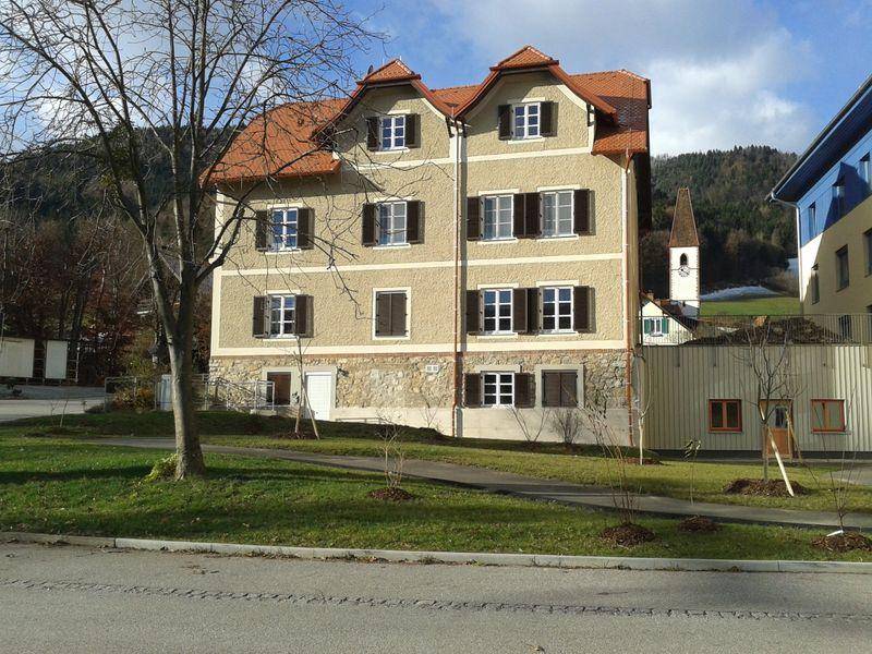 Gepflegtes Mehrfamilienhaus mit Satteldach und Gauben, umgeben von Grünflächen und Bergblick im Hintergrund.