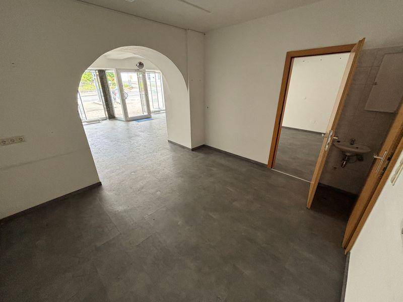Offener Geschäftsraum mit Blick zum Eingang und einem kleinen Waschbecken im angrenzenden Bereich.