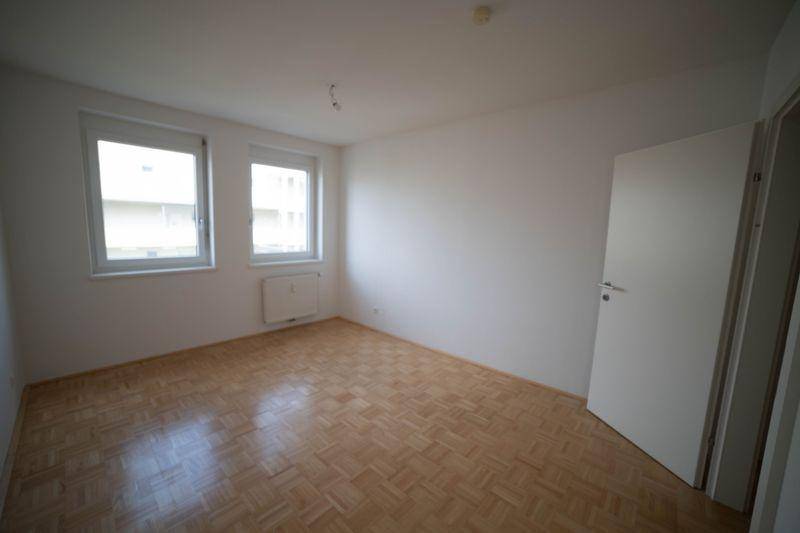 Helles, leeres Zimmer mit Parkettboden und zwei Fenstern, ideal als Schlafzimmer.