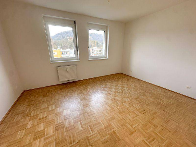 Leeres Schlafzimmer mit Parkettboden, zwei Fenstern und einem Heizkörper unter den Fenstern.