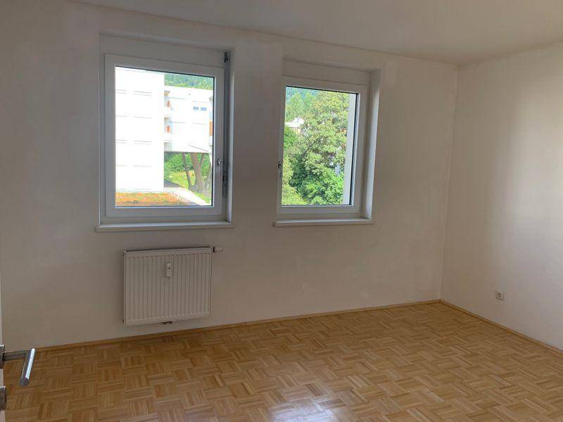 Leeres Zimmer mit Parkettboden, zwei Fenstern und Blick auf die grüne Umgebung.