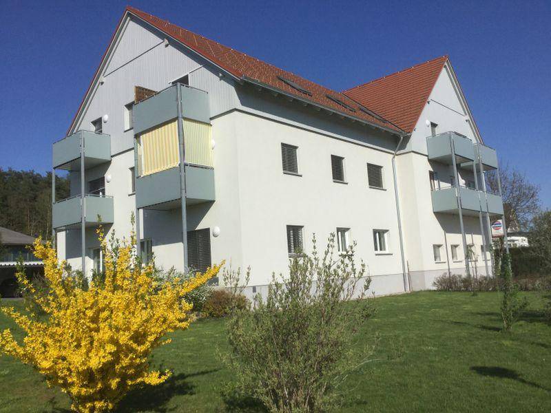 Modernes Mehrfamilienhaus mit Balkonen und rotem Ziegeldach, umgeben von grüner Landschaft.