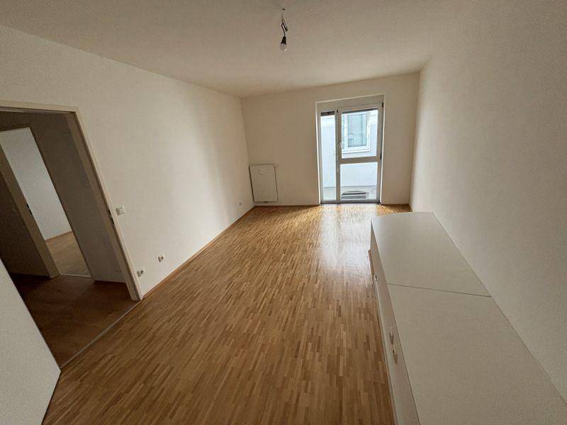 Geräumiges Wohnzimmer mit Holzfußboden, weißen Wänden und Zugang zum Balkon.