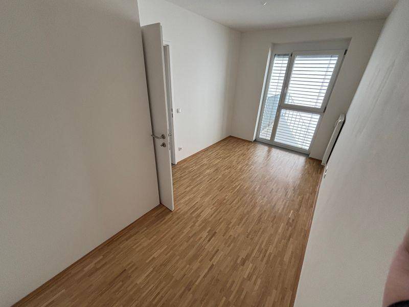 Leeres Zimmer mit hellem Parkettboden, weißen Wänden und großem Fenster mit Jalousien.