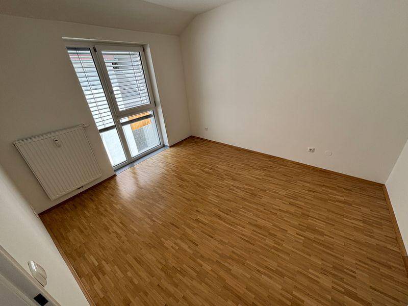 Helles Zimmer mit Parkettboden, weißen Wänden und großem Fenster mit Jalousien.