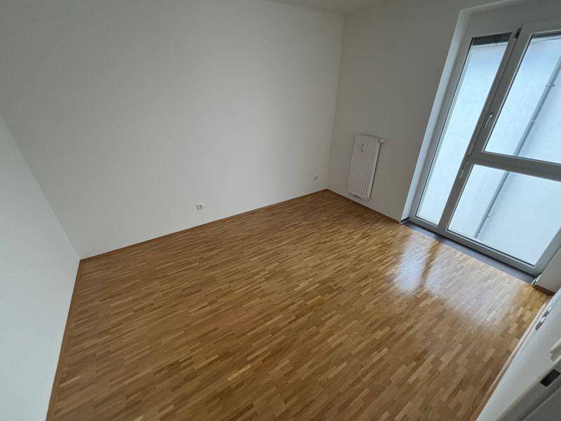 Leeres Zimmer mit hellem Parkettboden und großem Fenster, das viel Tageslicht hereinlässt.