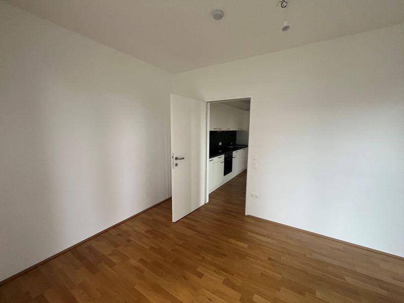 Leeres Wohnzimmer mit Holzboden und Zugang zu einer modernen Küche.