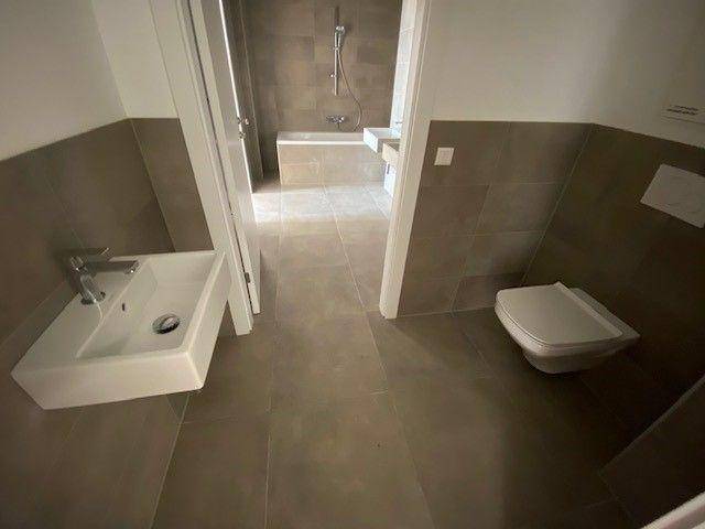 Modernes Badezimmer mit grauen Fliesen, separatem WC und Blick auf Badewanne und Waschbecken.