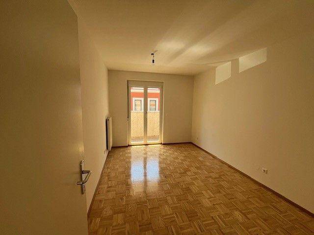Helles, leeres Zimmer mit Parkettboden und Zugang zu einem sonnigen Balkon.
