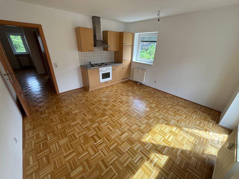 Wohnraum mit Parkettboden und integrierter Küchenzeile, hell durch Fenster und Balkontür.