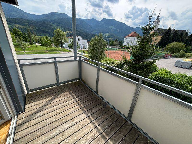 Geräumiger Balkon mit Holzboden und weitem Blick auf Berge, Kirche und Tennisplatz.