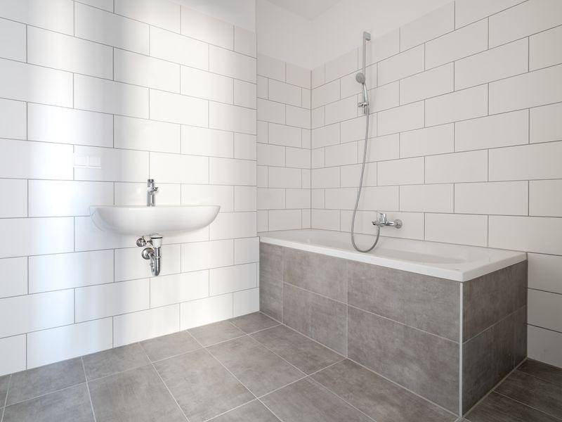 Helles Badezimmer mit weißen Wandfliesen, grauen Bodenfliesen, Badewanne mit Duschfunktion und Waschbecken.