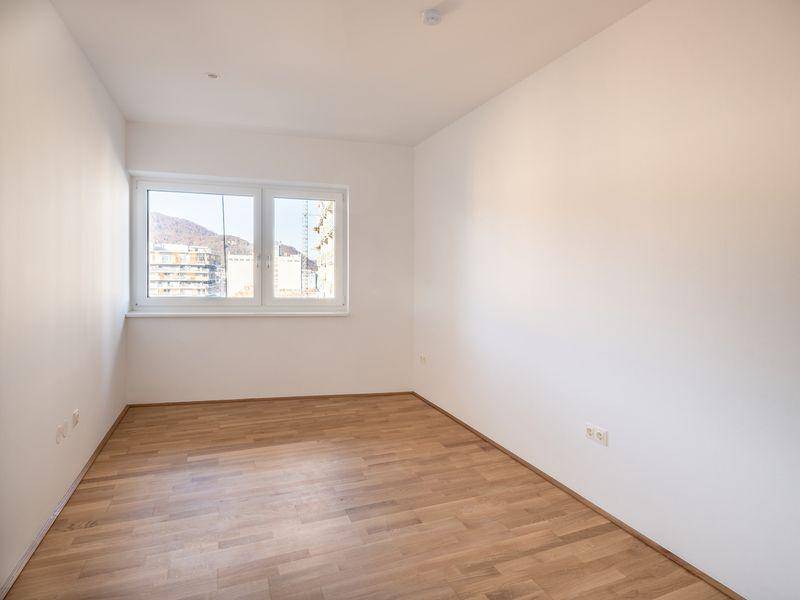 Leeres Zimmer mit hellem Holzfußboden und Fenster, das einen Ausblick auf die Umgebung bietet.