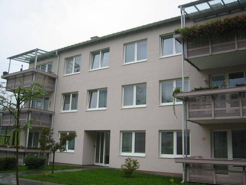 Mehrfamilienhaus mit heller Fassade, modernen Fenstern und mehreren Balkonen mit Glasüberdachung.