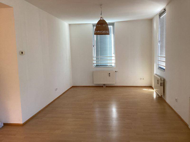 Geräumiges, leeres Wohnzimmer mit Laminatboden, Heizkörpern und großen Fenstern, die viel Licht hereinlassen.