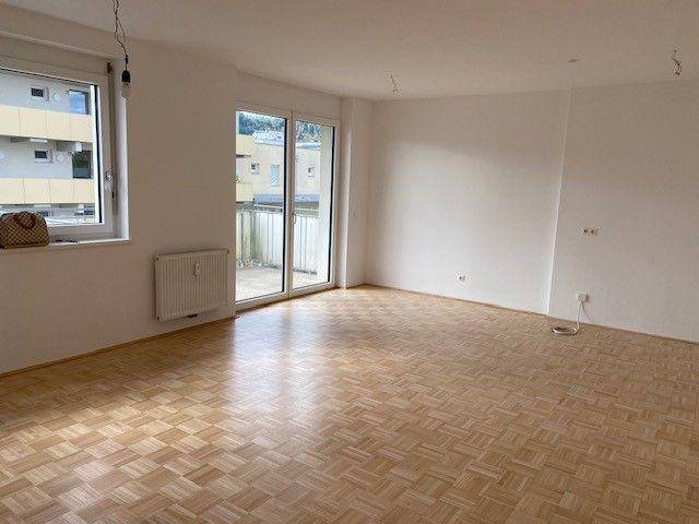 Geräumiges Wohnzimmer mit Parkettboden, großem Fenster und Zugang zu einem Balkon.