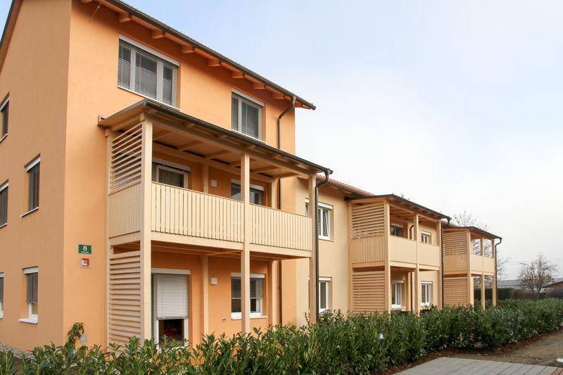 Modernes Mehrfamilienhaus mit orangefarbener Fassade, Holzverkleidung an den Balkonen und grüner Hecke.