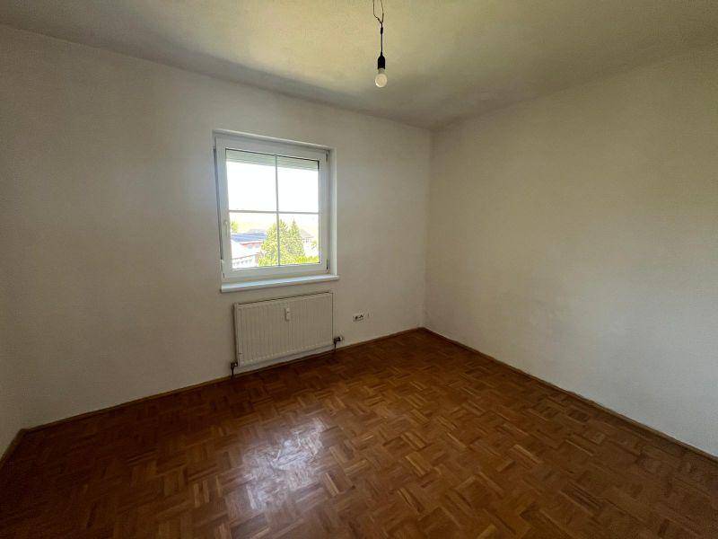 Leeres Zimmer mit Parkettboden und Fenster, das viel Tageslicht hereinlässt.