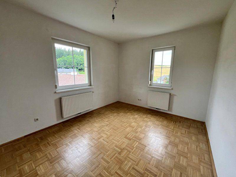 Geräumiges, leeres Zimmer mit zwei Fenstern, Parkettboden und Blick auf Bäume.