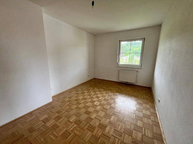 Leeres Zimmer mit Parkettboden, weiß gestrichenen Wänden und einem Fenster mit Blick ins Grüne.