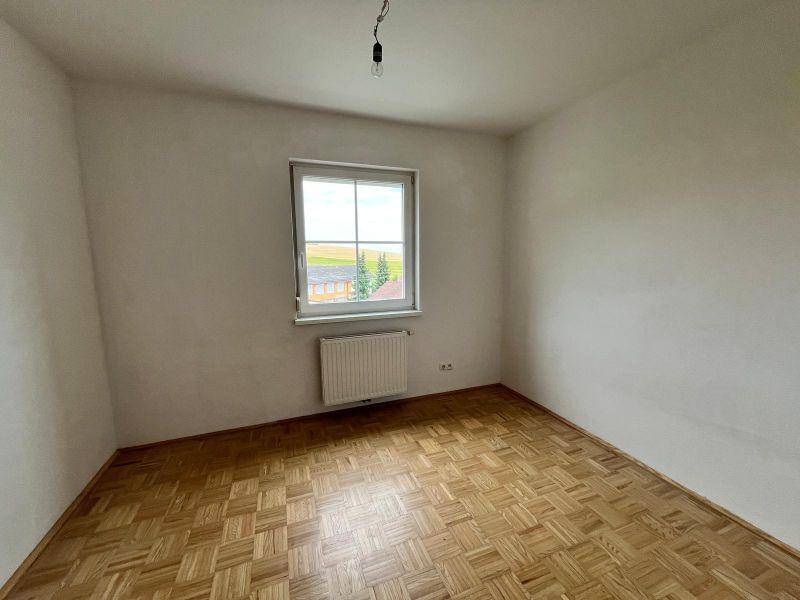 Helles, leeres Zimmer mit Parkettboden und einem Fenster, das einen Blick ins Grüne bietet.