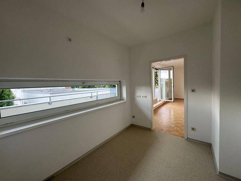 Helles Zimmer mit breitem Fenster und Zugang zu einem weiteren Raum mit Parkettboden.