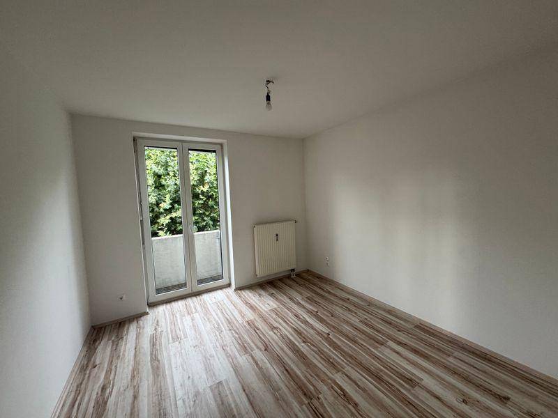 Geräumiges Zimmer mit Laminatboden und Zugang zu einem französischen Balkon mit Blick ins Grüne.