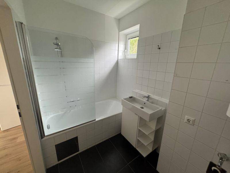 Helles Badezimmer mit Badewanne, Duschwand, Einzelwaschbecken und Fenster, ausgestattet mit weißen Fliesen.