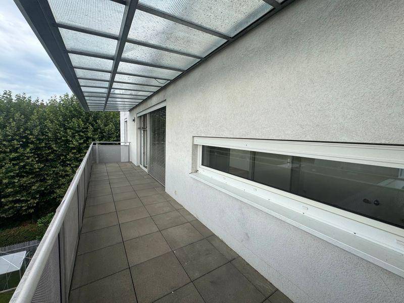 Langer, überdachter Balkon mit Fliesenboden und Metallgeländer, umgeben von grünen Bäumen.