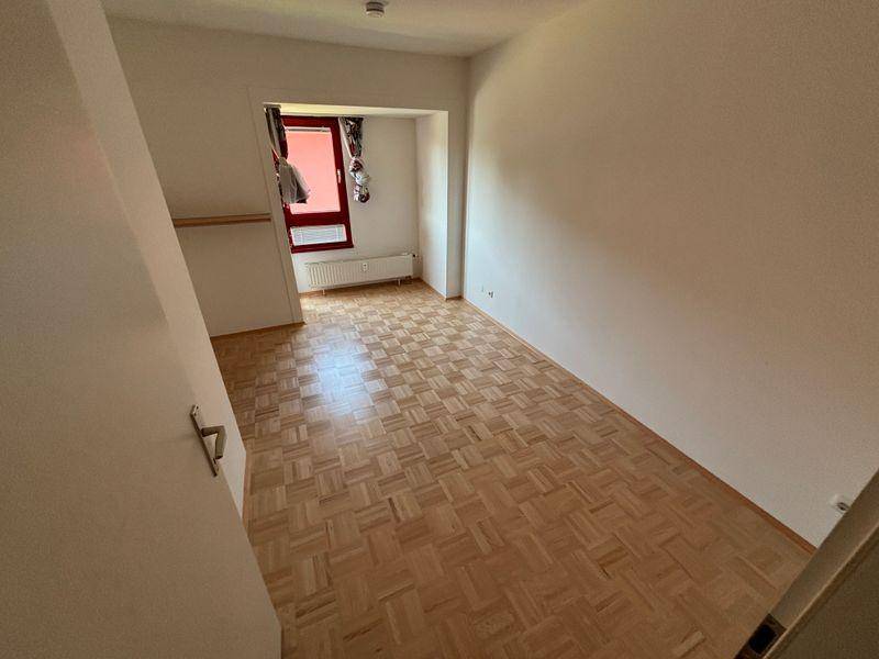 Leeres Schlafzimmer mit hellem Parkettboden und einem Fenster mit rotem Rahmen.
